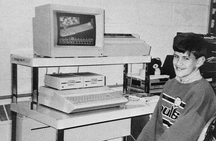 Black and white photograph of a student sitting in front of a computer workstation in 1990. The apple computer has a large keyboard built into the hardware assembly, a small CRT monitor, two large floppy disk drives, and a dot matrix printer. 