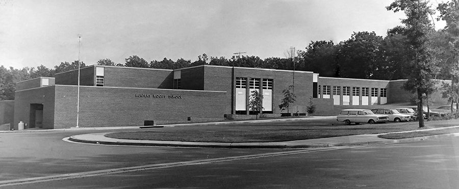 Black and white photograph of the front of Hunters Woods Elementary School taken in the early 1970s. The photographer is standing in the middle of Colts Neck Road looking up the hill toward the school. There are very few trees on the school grounds. A few small trees have been planted close to the main entrance.