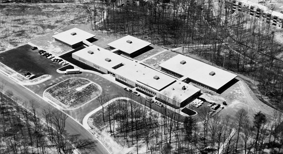 Black and white aerial photograph of Lake Anne Elementary School taken in the late 1960s or early 1970s. It is winter because all the leaves have fallen off the trees. The four classroom houses are distinctly visible. There is a school bus parked in front of the building and several cars are parked in the two small parking lots.