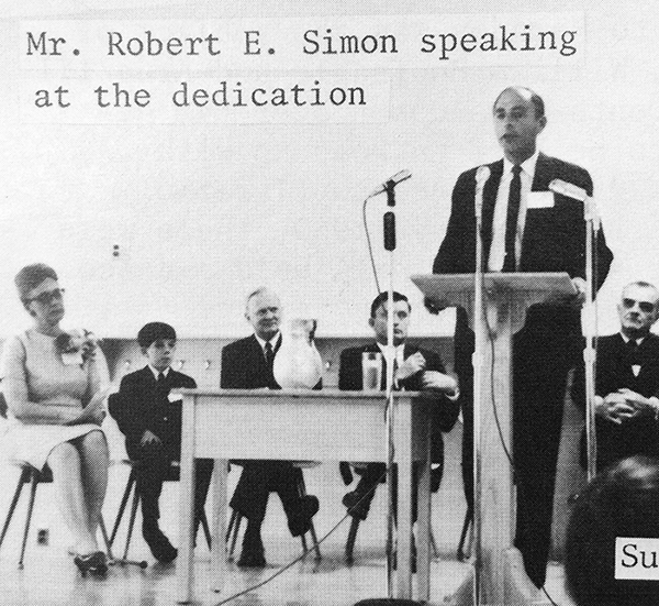 Black and white photograph of Robert E. Simon taken during the dedication ceremony of Lake Anne Elementary School. His is standing at a podium, surrounded by three microphones. Five people are seated behind him, one woman, one young boy, and three older men. The man on the far right is Superintendent Earl Funderburk.