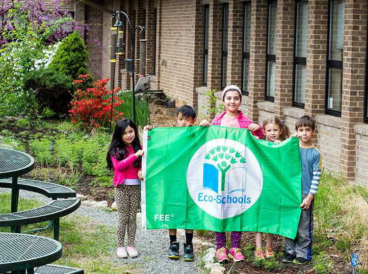 Photograph of five students holding up the Eco-Schools green flag. They are standing outside Lake Anne Elementary School in the courtyard garden. The picture was taken in April 2015, and behind the students flowers and trees are blooming with red, white, and purple blossoms. The garden is lush with new green growth.