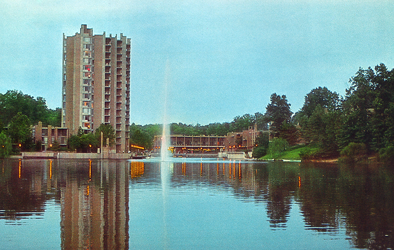 Color photograph of Reston's Lake Anne Village Center taken from a boat on Lake Anne looking toward the high-rise condominium tower and the shopping center. 