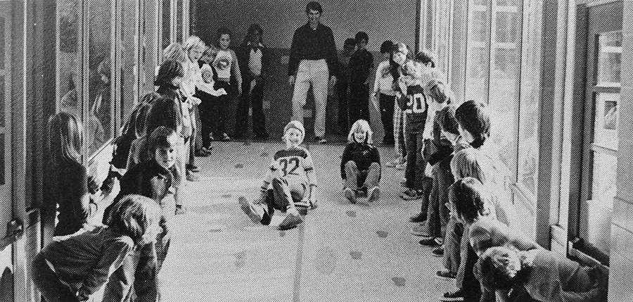 Black and white photograph of a physical education class from our 1975 to 1976 yearbook. The students are lined up along the walls in a hallway cheering on two students who are racing each other crab-walking on sliding scooters. 