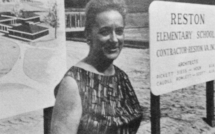 Black and white photograph of Principal Ward taken in 1966. She is standing in front of Lake Anne Elementary School, which is still under construction. Two signs have been placed in front of the school. One is the concept artwork shown earlier on this webpage, and the second sign has text that reads: Reston Elementary School, and gives the name of the contractor and architects. 