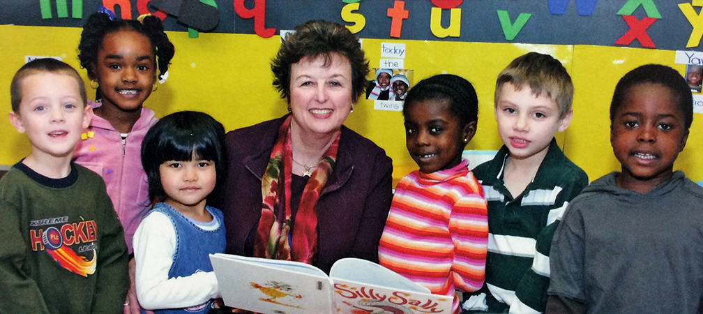 Yearbook photograph of Principal Linda Hajj taken in 2008. She is holding a picture book entitled Silly Sally in her hand. Three children stand on her right, and three children stand on her left. Everyone is looking at the camera and smiling.