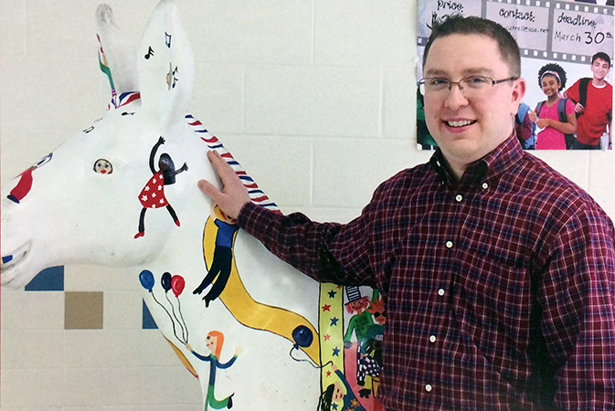 Yearbook photograph of Principal Brendan Menuey taken in 2014. He is standing in the school lobby with his hand on the Party Animal donkey statue. 