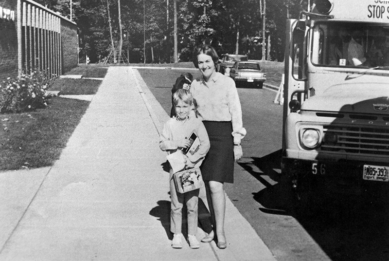 Black and white photograph of Principal Margie Thompson and an unnamed male student from our 1969 to 1970 yearbook. They are standing on the sidewalk in front of Lake Anne Elementary School next to a school bus that is parked with its door open. The boy is holding a lunch box and has an armful of books and papers. 