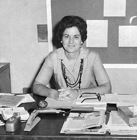 Black and white photograph of Principal Margie Thompson from our 1970 to 1971 yearbook. She is seataed at her desk, looking up from a large stack of paperwork. 